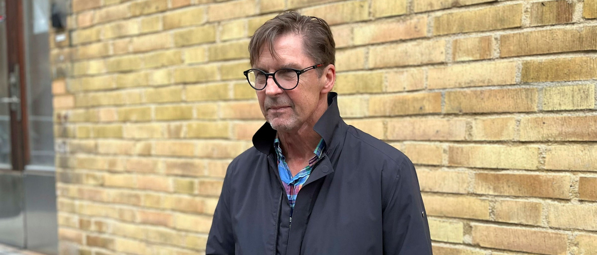  Per-Anders står framför en gul tegelvägg, han bär svarta glasögon och en rock.