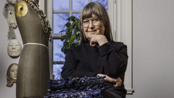 Monica Hallen sitter bakom en trave med blå trasvävar. till vänster syns en provdocka med smycken och i bakgrunden ett spröjsat fönster och masker på en vägg.