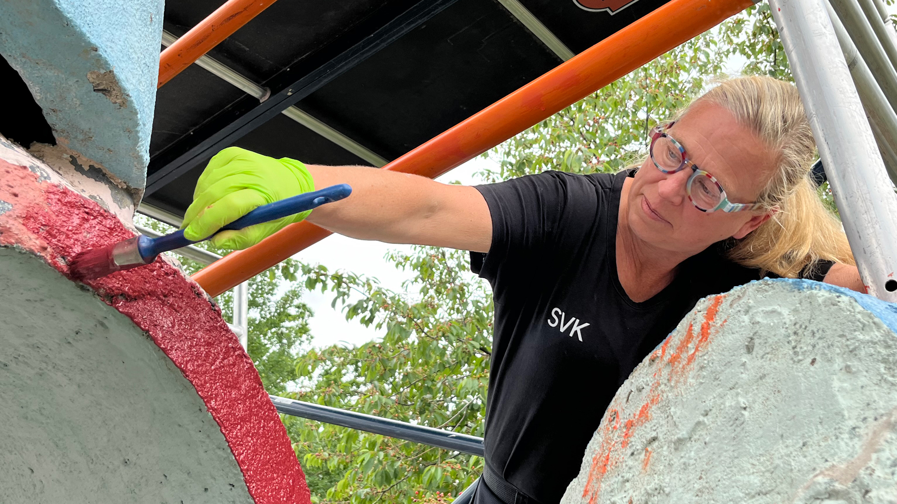 Kvinna, blond i svart t-shirt, målar. Oklart vad, men hon lutar sig framåt. Orange stålrör på bilden. 