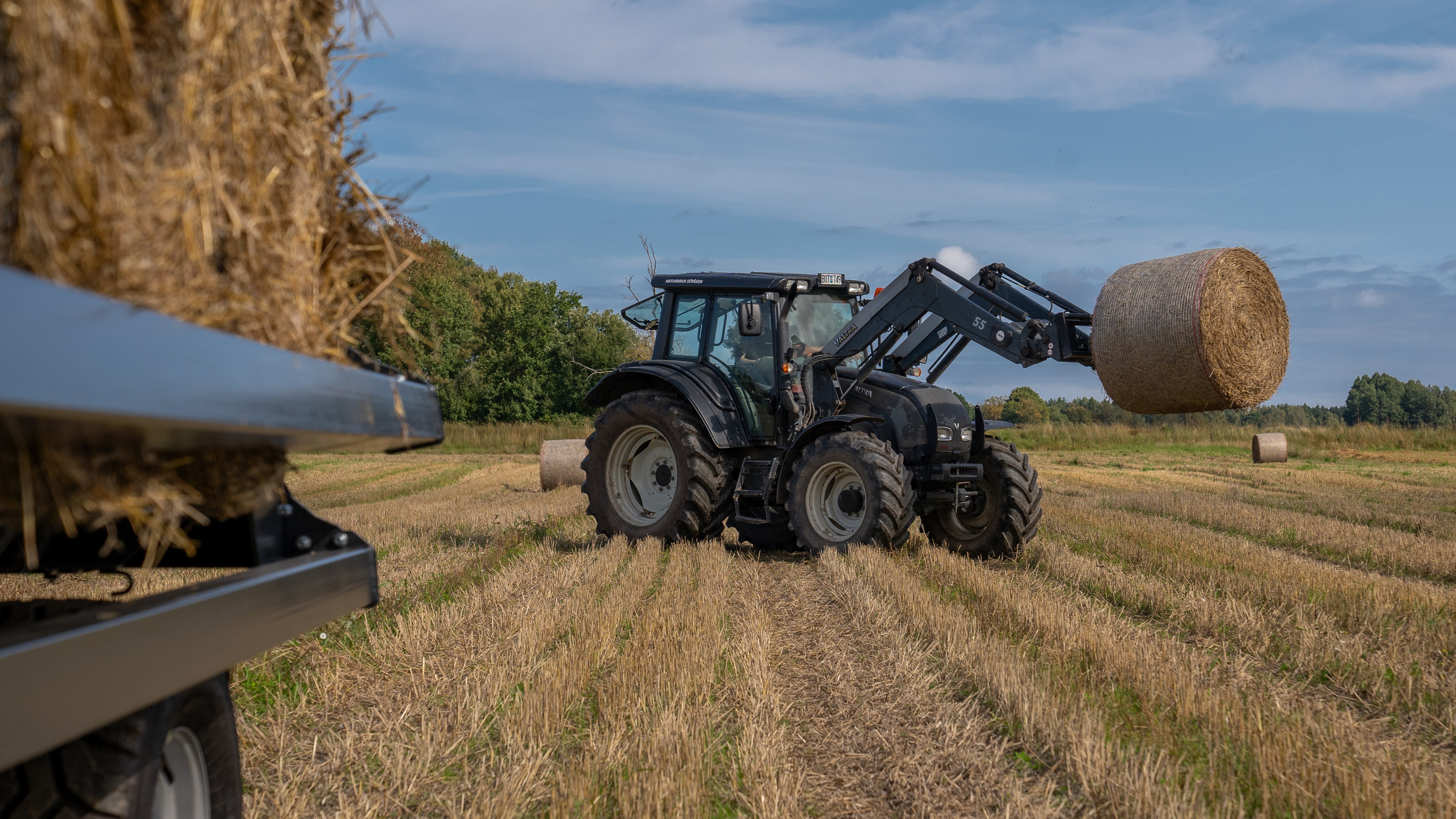 Traktor lyfter upp halmbal med balvagn i förgrunden