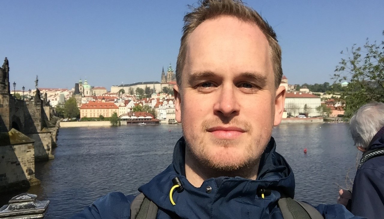 Man i 30-års åldern. Blå jacka, stad i bakgrunden samt vattendrag.
